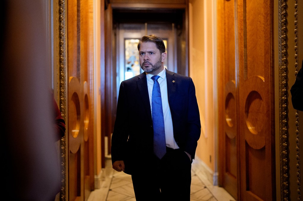 Sen. Ruben Gallego walks through a wood-paneled hallway in a dark suit and blue tie.