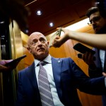 Sen. Mark Kelly stands in an elevator doorway as reporters hold phones and recording devices toward him.