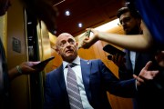 Sen. Mark Kelly stands in an elevator doorway as reporters hold phones and recording devices toward him.