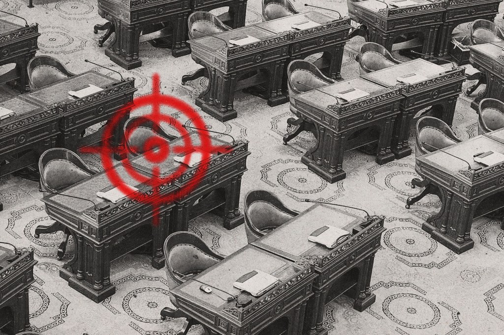 Black and white photo of an empty legislative chamber with rows of wooden desks and chairs. A bright red, spray-painted target symbol is scrawled across one desk in the foreground.