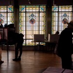 People vote at individual booths inside a dimly lit polling place, silhouetted against large stained-glass windows in the background.