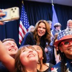 A group of people flank a woman on stage with American flags in the background.