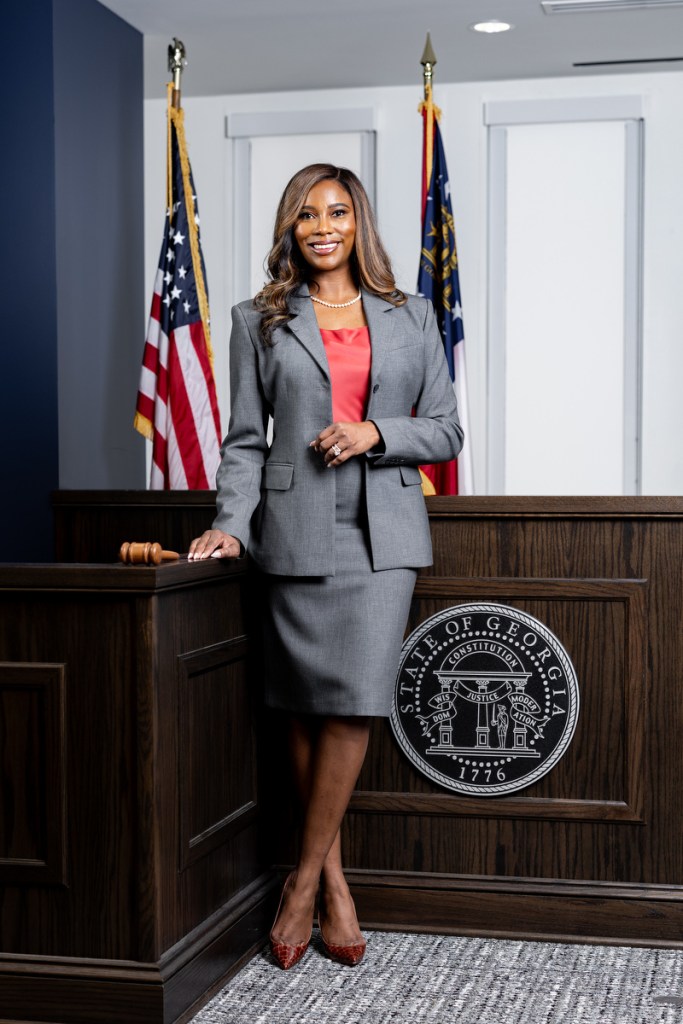 A portrait of a woman standing in a courthouse flanked by American flags and the seal of Georgia behind her.