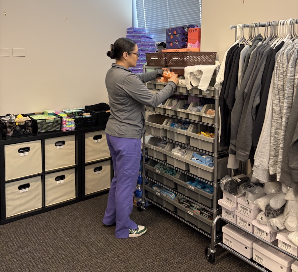 A person wearing purple scrub pants and a gray jacket organizes medical supplies on a metal shelving unit in a small storage room. Bins filled with items like gloves, bottles, and packaged materials line the shelves, while clothing hangs on a rack nearby.