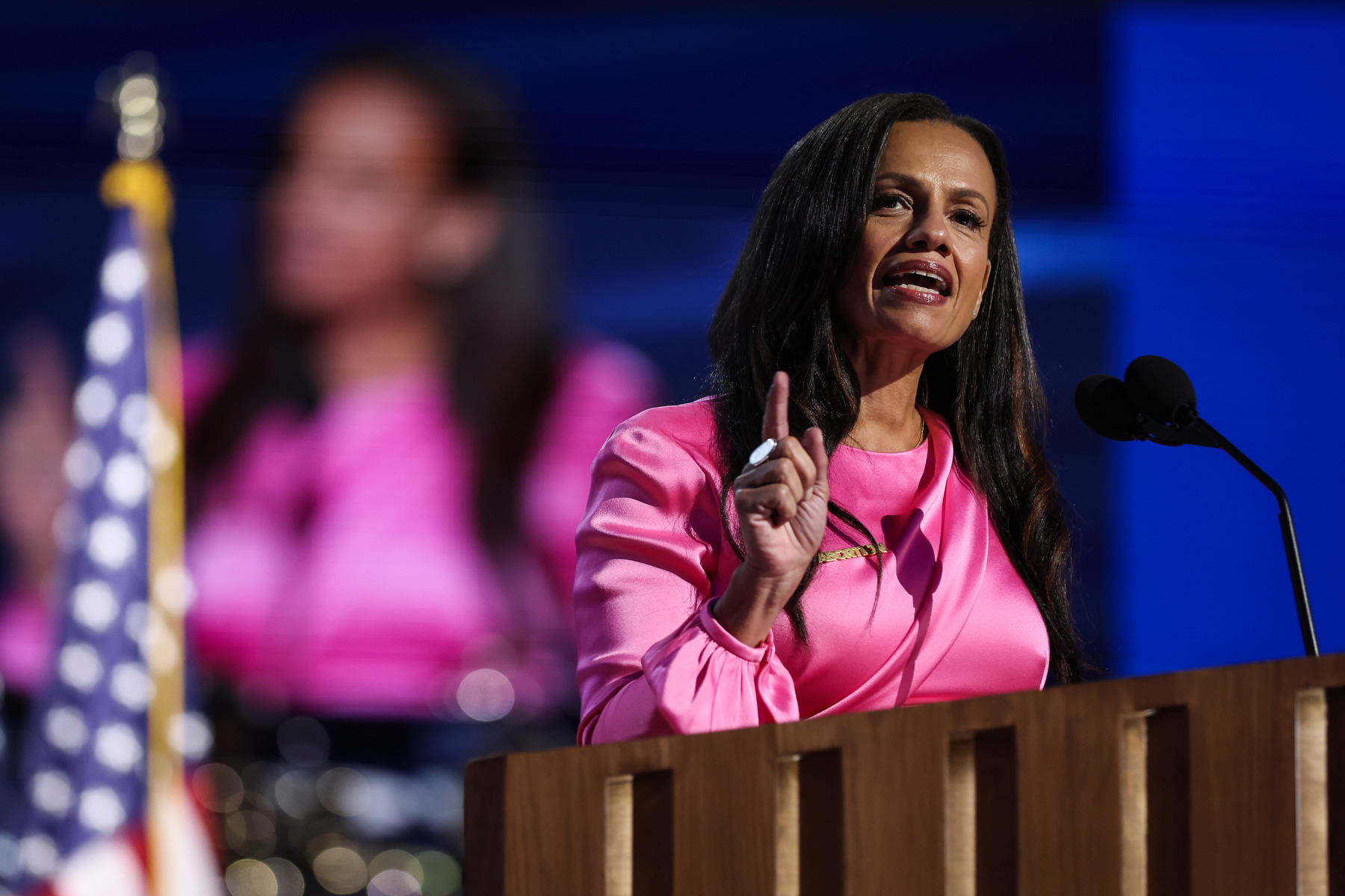 A woman in pink speaks at a podium, pointing her finger.