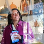A woman in a pink coat smiles while standing inside a shop, holding a stack of flyers and a coffee. Reflections in the glass partially overlay her face, and shelves behind her display decorative objects.
