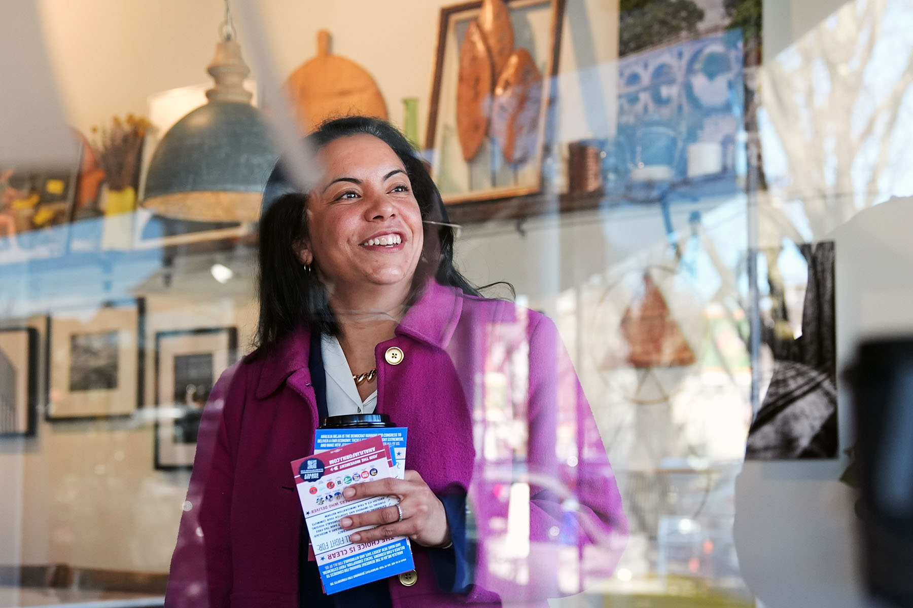 A woman in a pink coat smiles while standing inside a shop, holding a stack of flyers and a coffee. Reflections in the glass partially overlay her face, and shelves behind her display decorative objects.