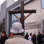 A person holds a large wooden cross while standing outside a Planned Parenthood clinic, where a group of people gather on the sidewalk in winter clothing.