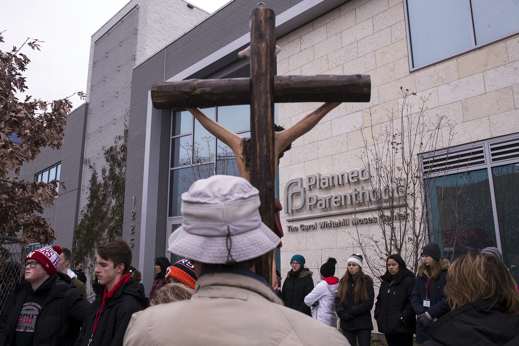 A person holds a large wooden cross while standing outside a Planned Parenthood clinic, where a group of people gather on the sidewalk in winter clothing.
