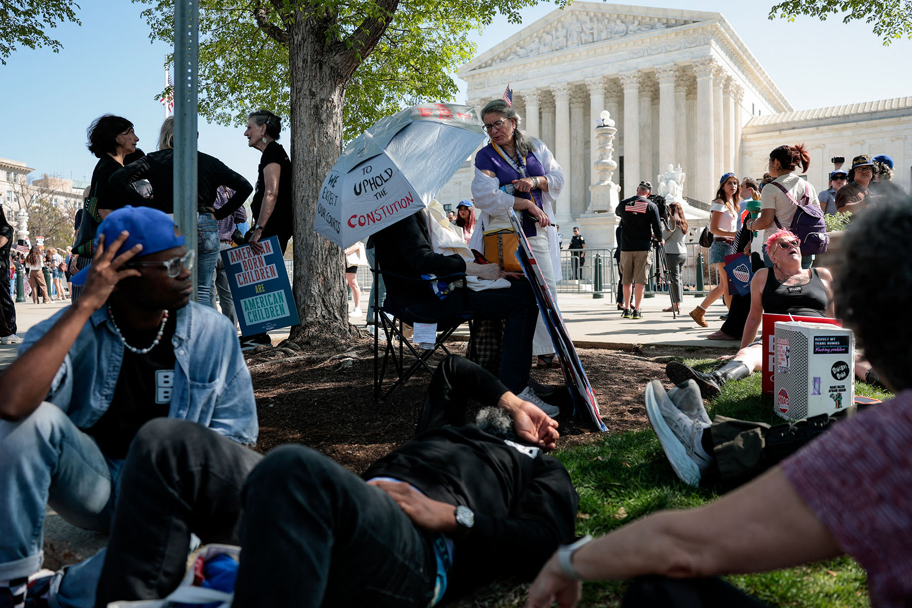 People gather outside the U.S. Supreme Court in Washington, D.C., with demonstrators resting in the shade and holding signs about birthright citizenship and the Constitution.
