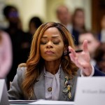 Rep. Sheila Cherfilus-McCormick sits at a hearing table with her eyes closed during a House hearing. She wears a gray blazer with a large bird brooch and pink earrings, while blurred audience members and another speaker’s gesturing hands appear in the background.