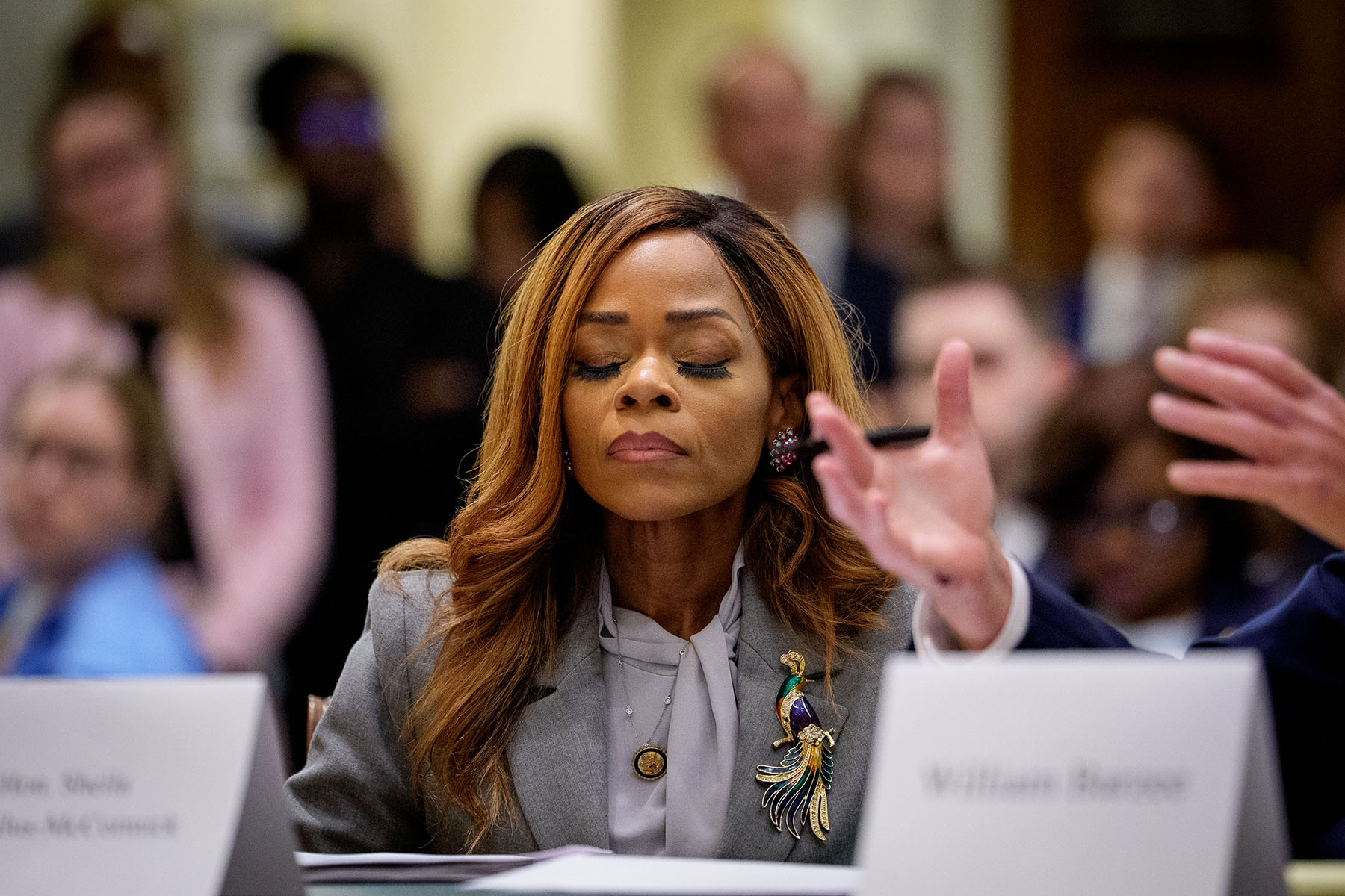 Rep. Sheila Cherfilus-McCormick sits at a hearing table with her eyes closed during a House hearing. She wears a gray blazer with a large bird brooch and pink earrings, while blurred audience members and another speaker’s gesturing hands appear in the background.