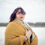 A person with purple hair looks at the camera in a portrait against a snowy landscape.