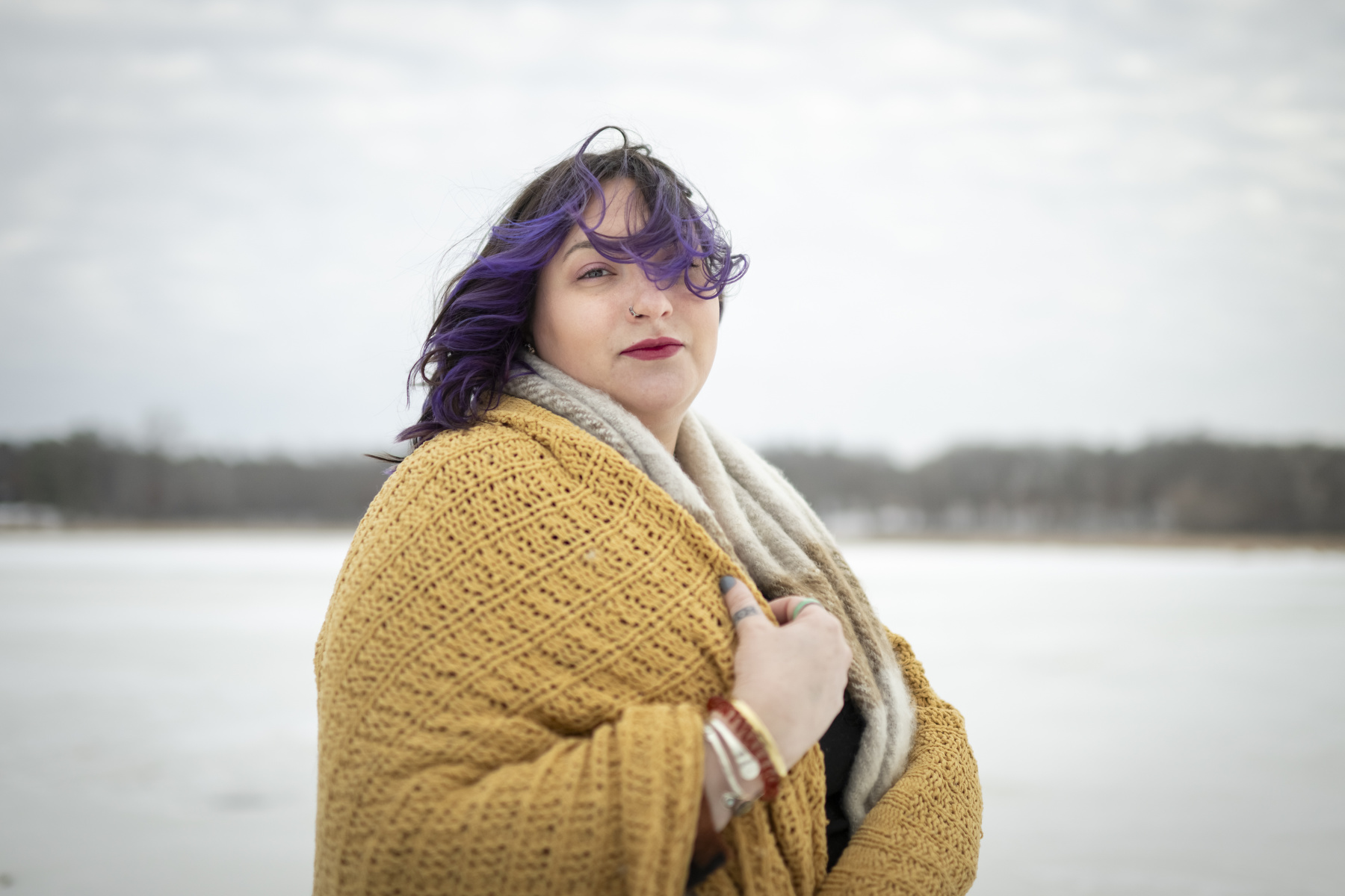 A person with purple hair looks at the camera in a portrait against a snowy landscape.