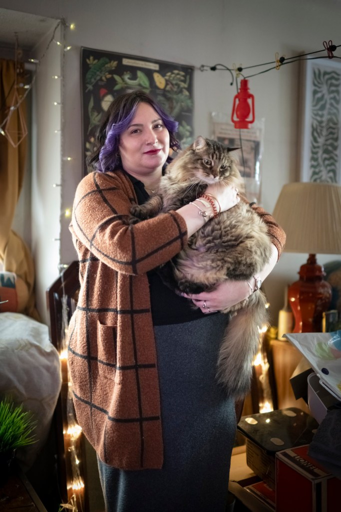 A person with purple hair looks at the camera in a portrait while holding a cat in a living room.