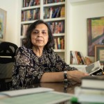 A woman sits at a desk in her home looking into the camera.
