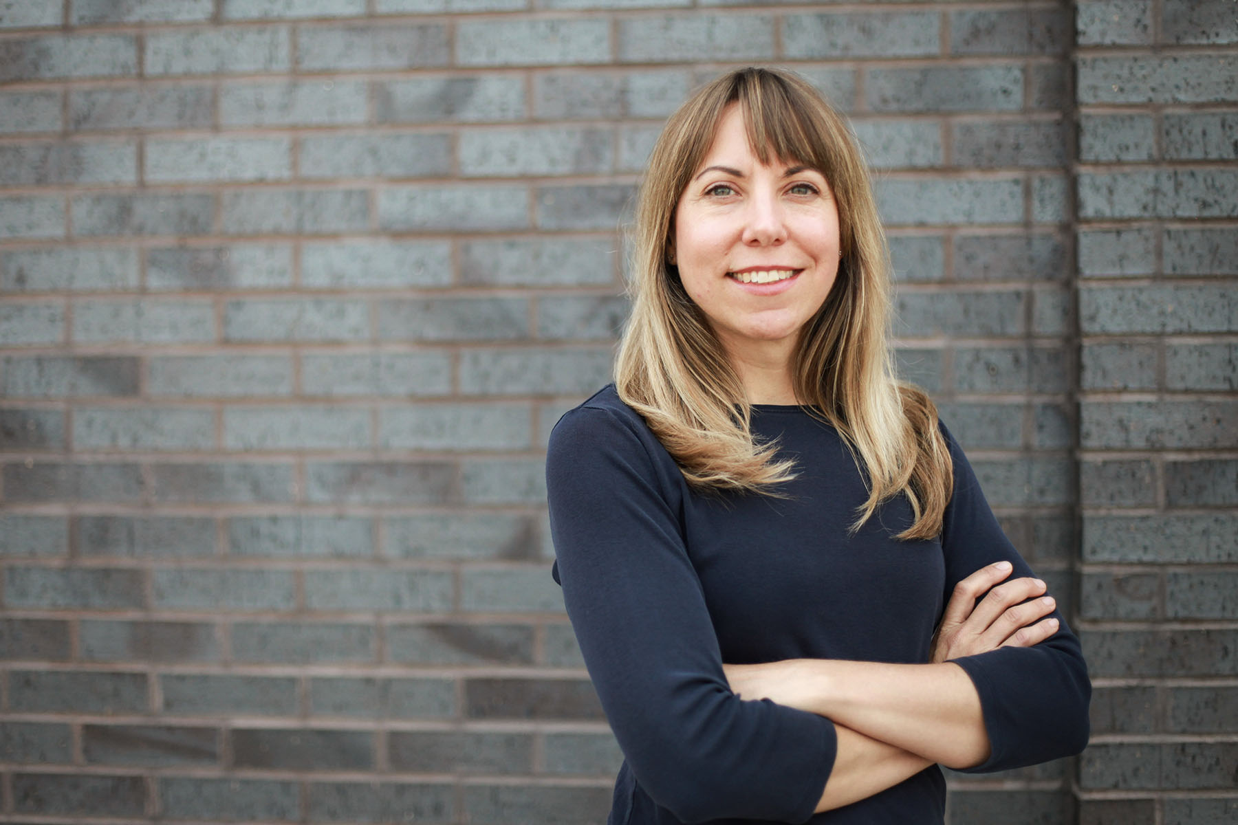 A woman with shoulder-length light brown hair stands with her arms crossed in front of a gray brick wall looking at the camera.