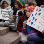 Several children sit on stone steps at a protest, wearing colorful winter clothing and holding handmade signs, including one that reads “Let Trans Kids Be” and another that says “Gender Liberation Now.”