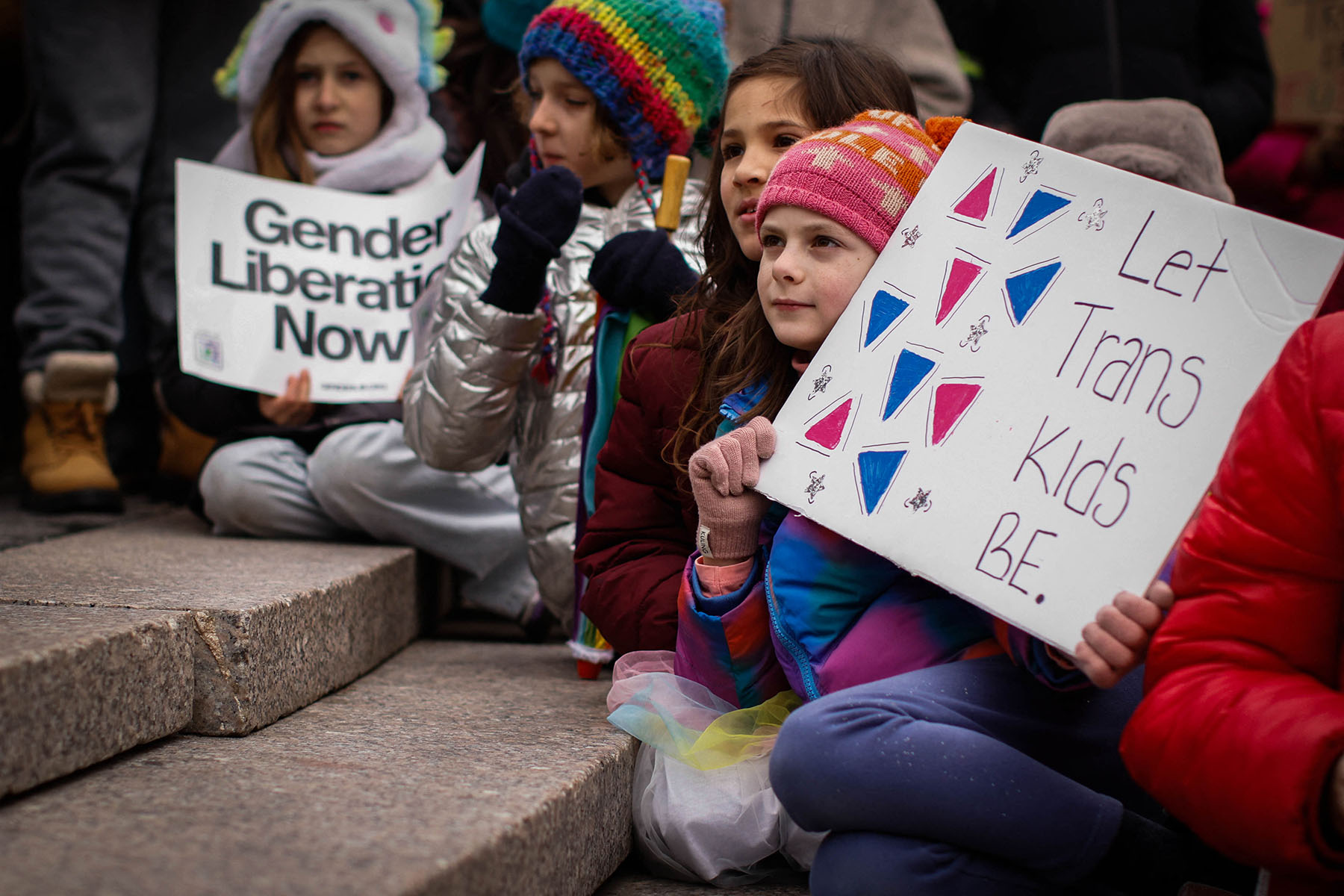 Several children sit on stone steps at a protest, wearing colorful winter clothing and holding handmade signs, including one that reads “Let Trans Kids Be” and another that says “Gender Liberation Now.”