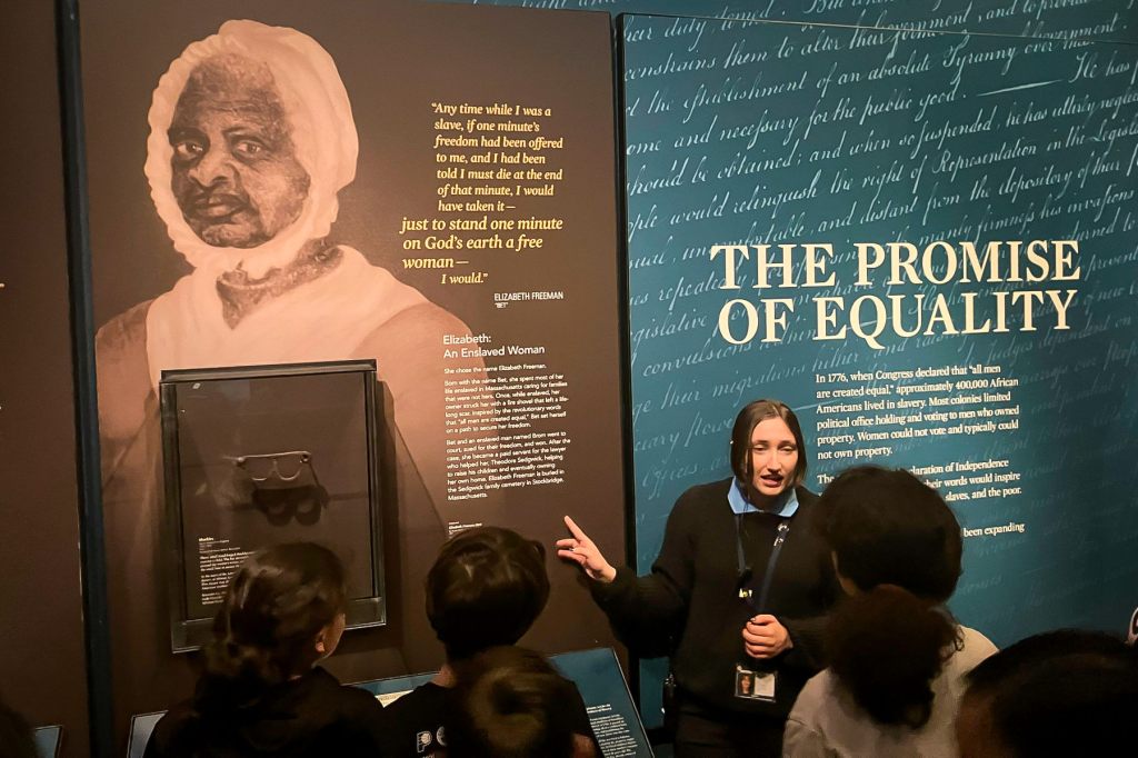 A guide, speaking to a group of children, points to a wall mural depicting a formerly enslaved woman and the words “the promise of equality.”