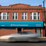 Exterior of a brick building with a teal awning labeled “Planned Parenthood.” A blurred car passes in front of the clinic on the street.