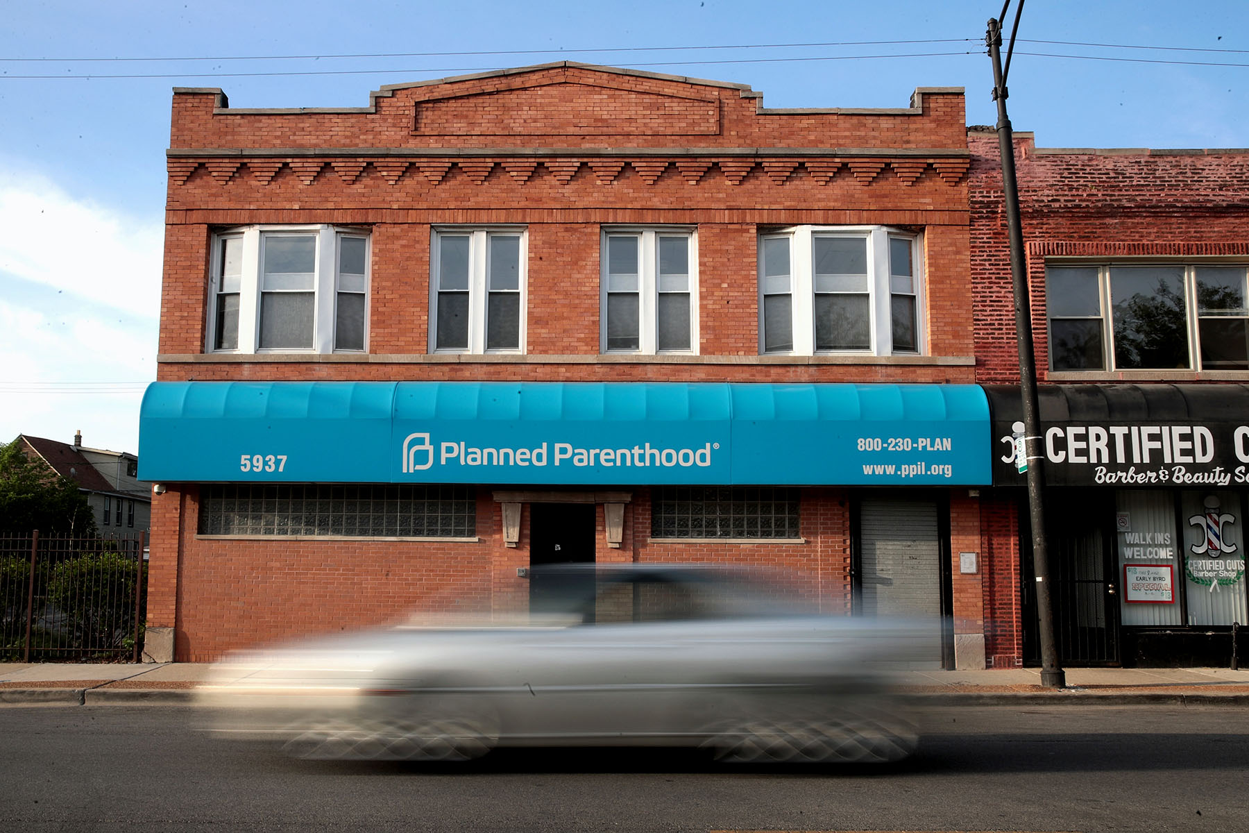Exterior of a brick building with a teal awning labeled “Planned Parenthood.” A blurred car passes in front of the clinic on the street.