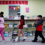 A line of students hold books while inside a library. A wall in the back bears a sign that says Everybody.
