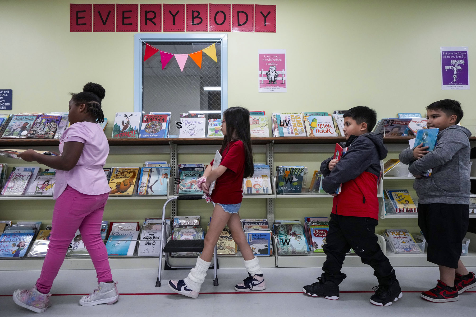 A line of students hold books while inside a library. A wall in the back bears a sign that says Everybody.