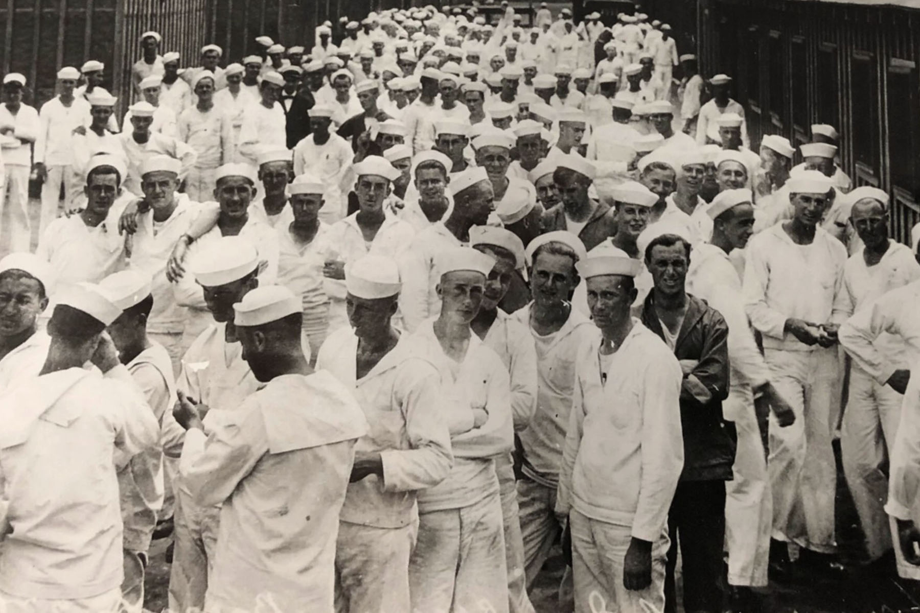 A large group of sailors in white uniforms and caps stand closely together outdoors, filling the frame as they face the camera.