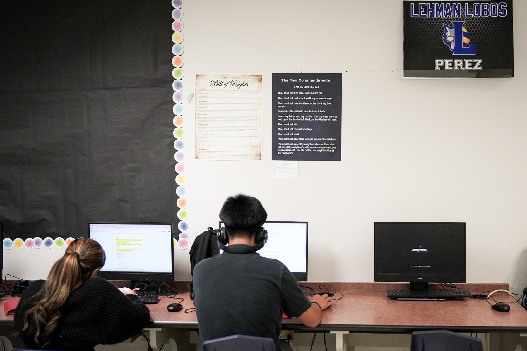 Two students work at computers with posters of the Bill of Rights and Ten Commandments hanging above their heads.