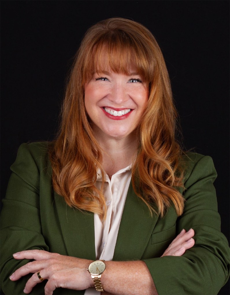 A woman with long red hair smiles at the camera against a dark background, with her arms crossed.