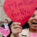 A domestic worker holds a large red paper heart during a City Hall advocacy action in New York City while Allison Julien stands in the background with a pink paper heart.