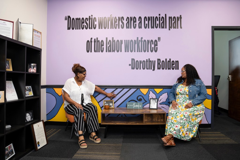 Vernell Truitt and Alicia Cleveland sit in the National Domestic Workers Alliance office beneath a wall quote from Dorothy Bolden that reads, "Domestic workers are a crucial part of the labor workforce."