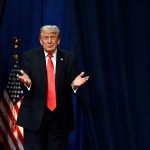 Trump wears a dark suit and red tie as he stands with his hands raised in front of dark blue curtains, with an American flag partly visible behind him.