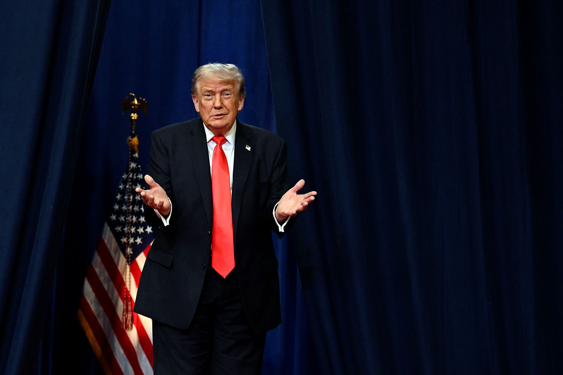Trump wears a dark suit and red tie as he stands with his hands raised in front of dark blue curtains, with an American flag partly visible behind him.