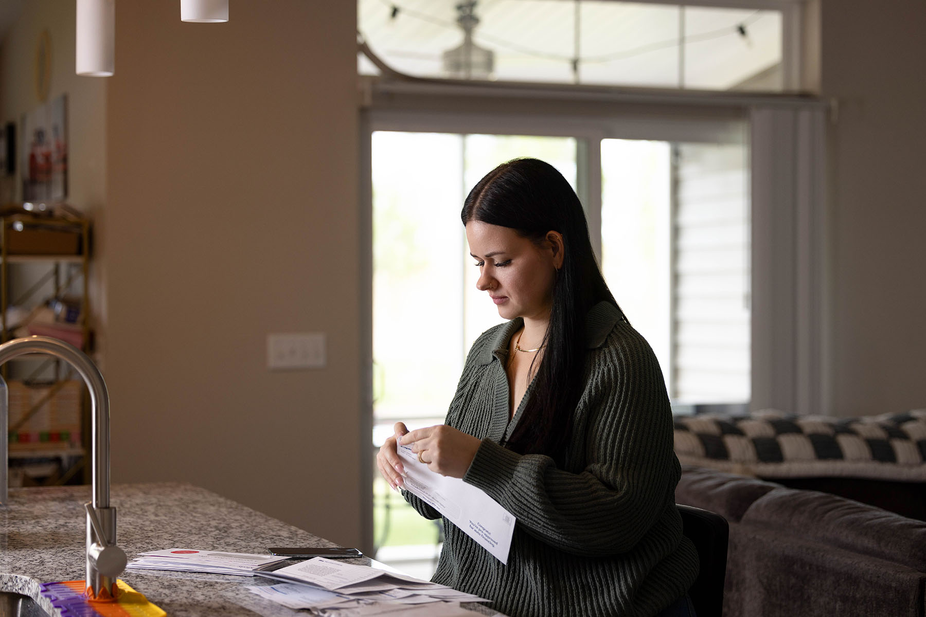 Harmonie Perrone sits at a kitchen counter, opening mail beside a stack of envelopes and papers.