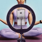 A person in lavender workout clothes sits cross-legged on a yoga mat behind a ring light and smartphone, with their arms extended in a meditation pose.