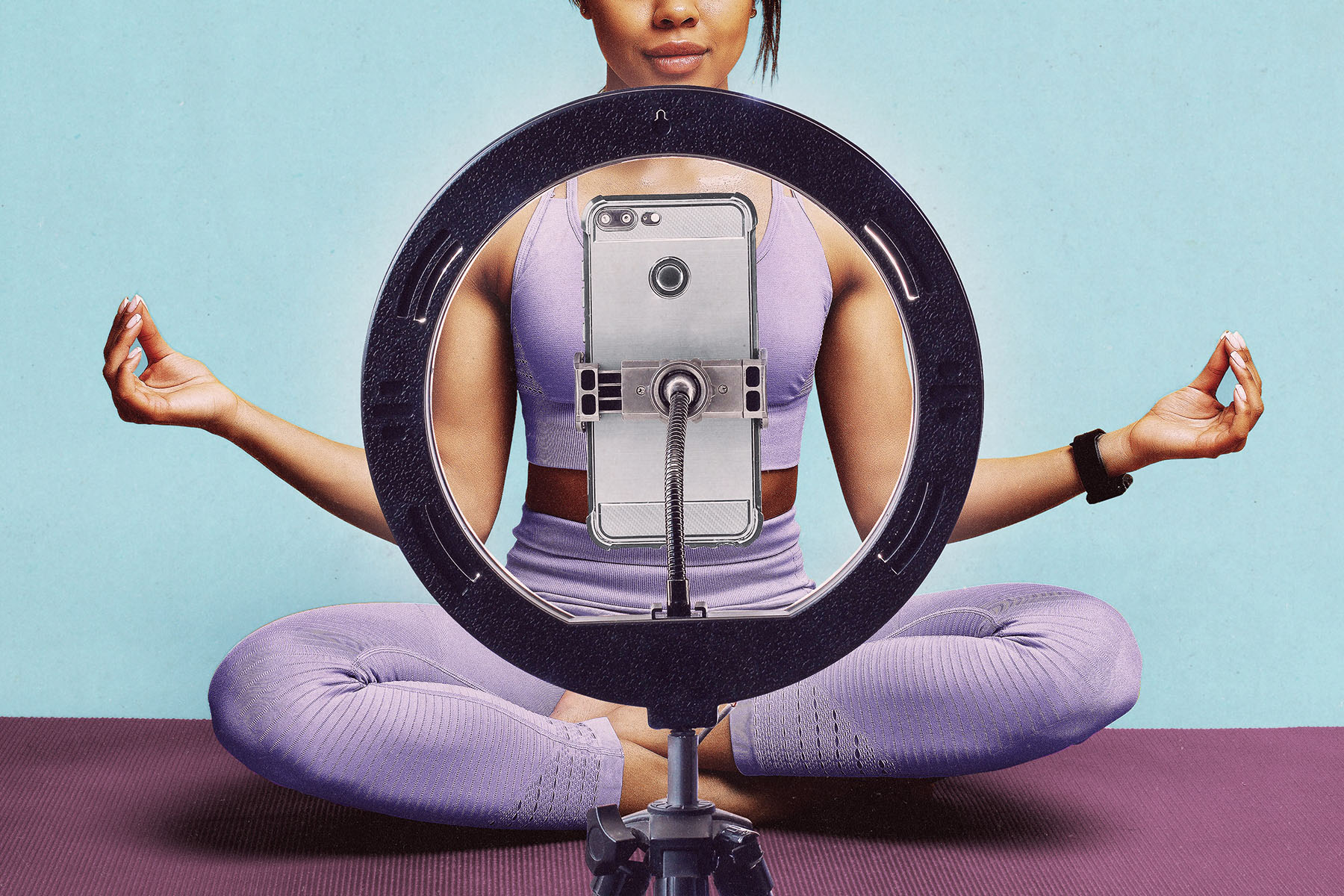 A person in lavender workout clothes sits cross-legged on a yoga mat behind a ring light and smartphone, with their arms extended in a meditation pose.