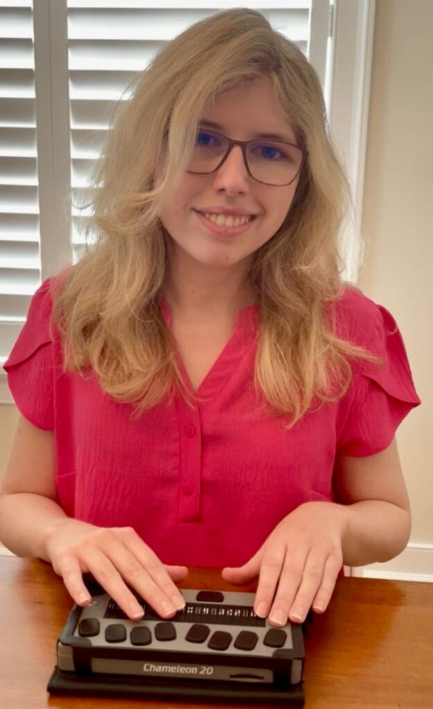 A young woman with blond hair and glasses smiles while seated at a table with her hands on a Chameleon 20 braille display.