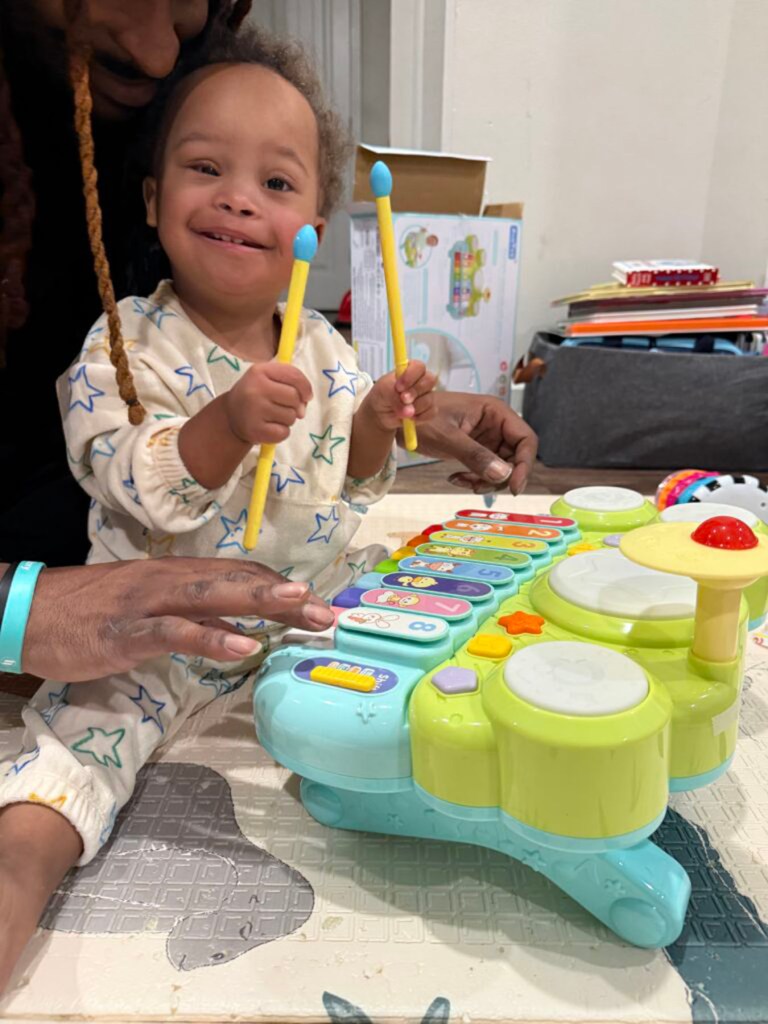 A toddler smiles while holding two toy drumsticks above a colorful toy drum set, with an adult seated behind them.