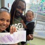 A family takes a selfie indoors, holding a drawing that says “Landry” in purple crayon.