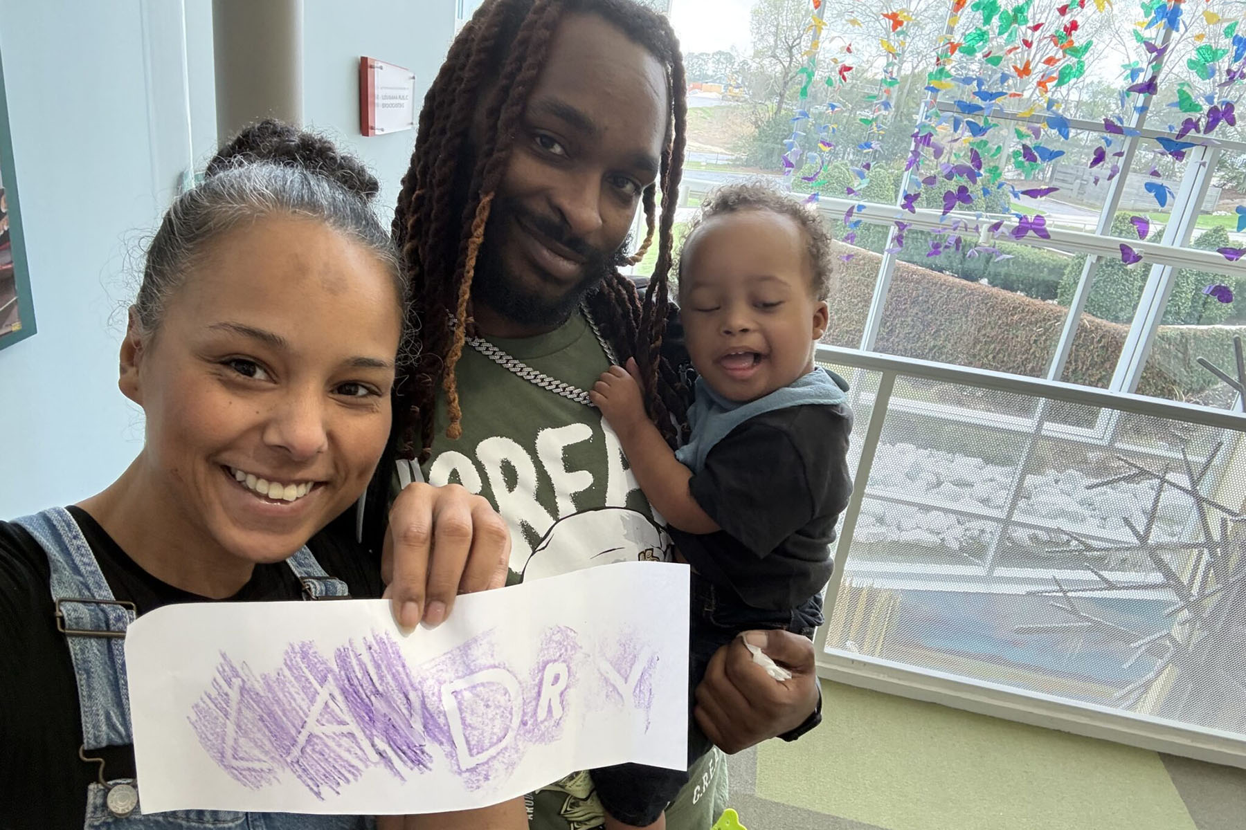 A family takes a selfie indoors, holding a drawing that says “Landry” in purple crayon.
