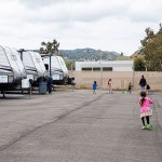 Children play on scooters in a paved lot lined with travel trailers.