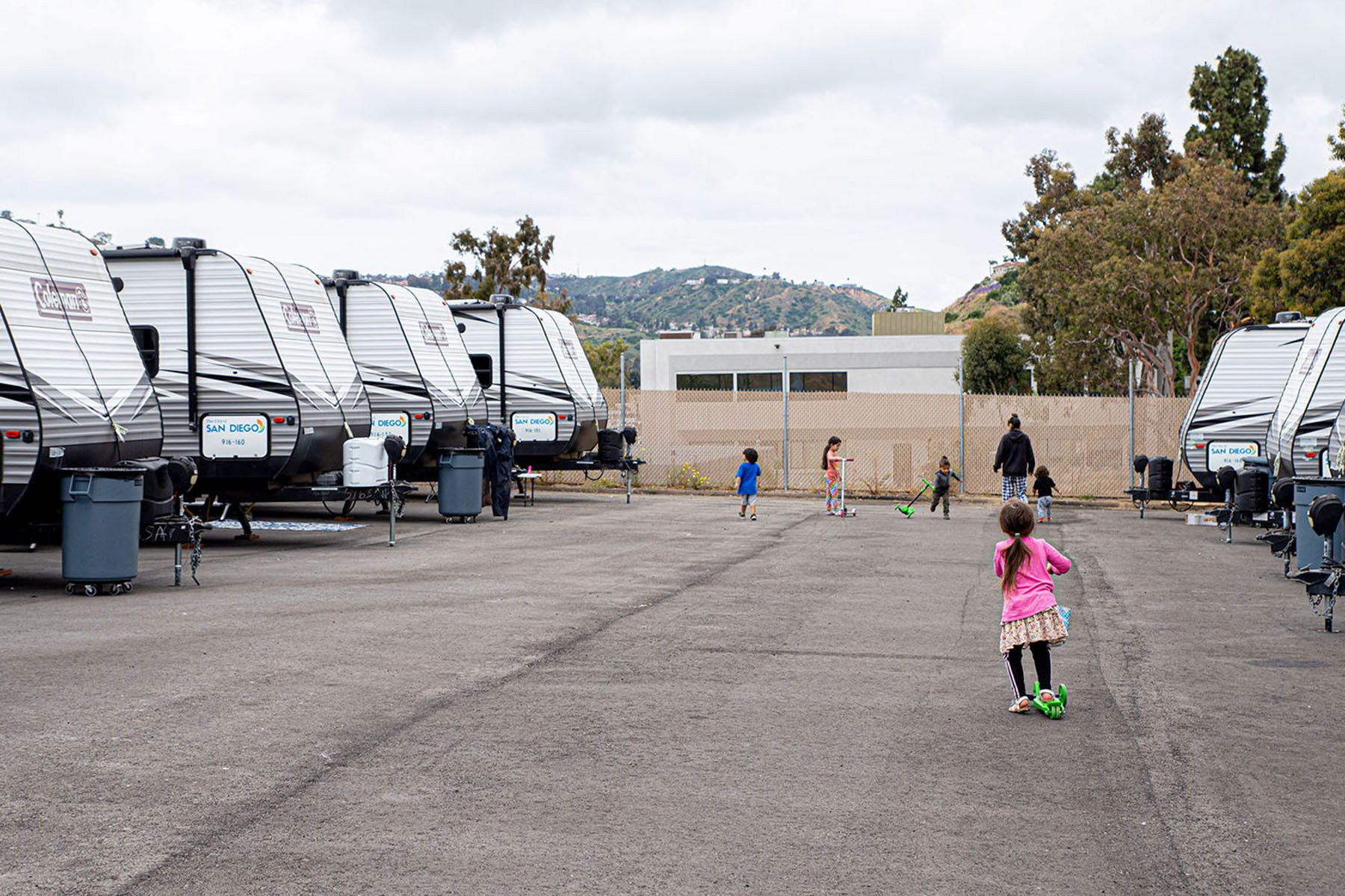 Children play on scooters in a paved lot lined with travel trailers.