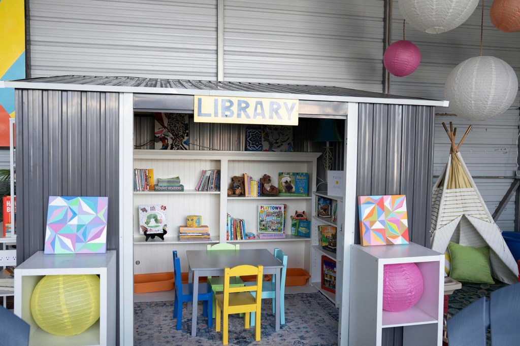 A children’s library area with bookshelves, small chairs, paper lanterns and a teepee inside a shared indoor space.