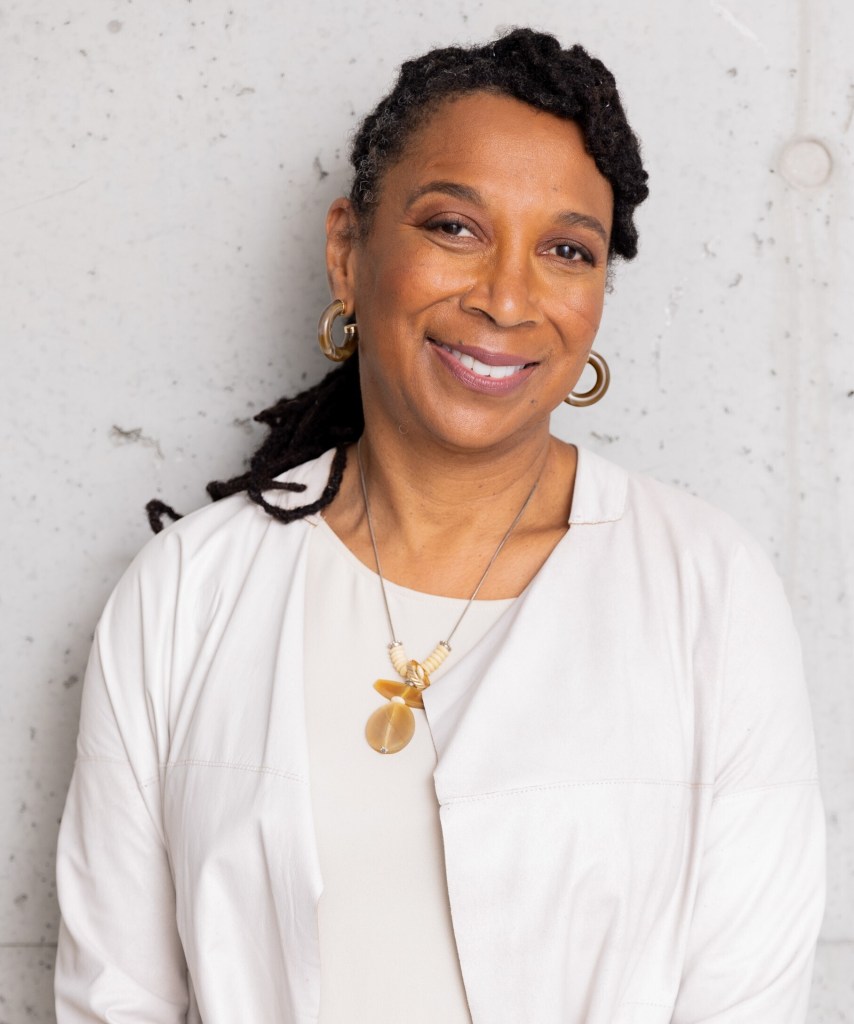 Kimberlé Crenshaw smiles at the camera in a portrait against a wall, wearing a white top, gold hoop earrings and a necklace.