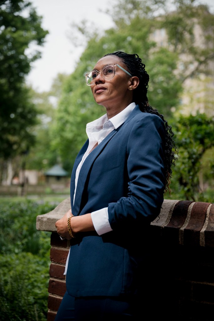 Adriana George stands outside leaning against a low brick wall in a navy suit and white button down shirt.