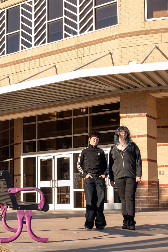 Two students stand outside the entrance of a brick school building near a pink bench.