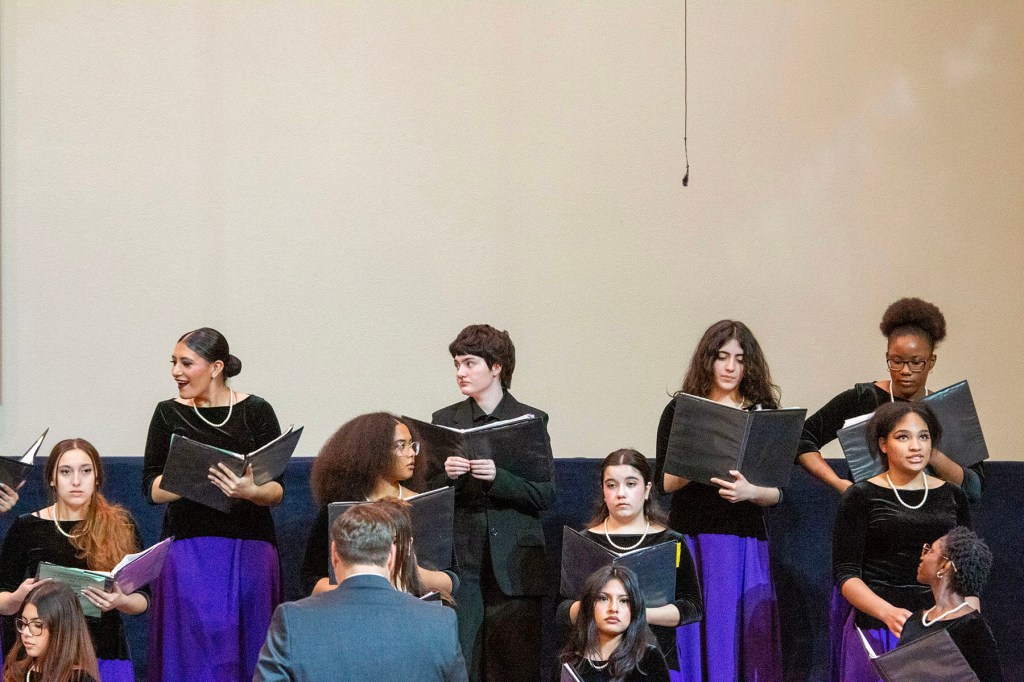 A student choir performs indoors, holding black folders.