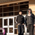 Two students stand outside a brick school building in warm sunlight.